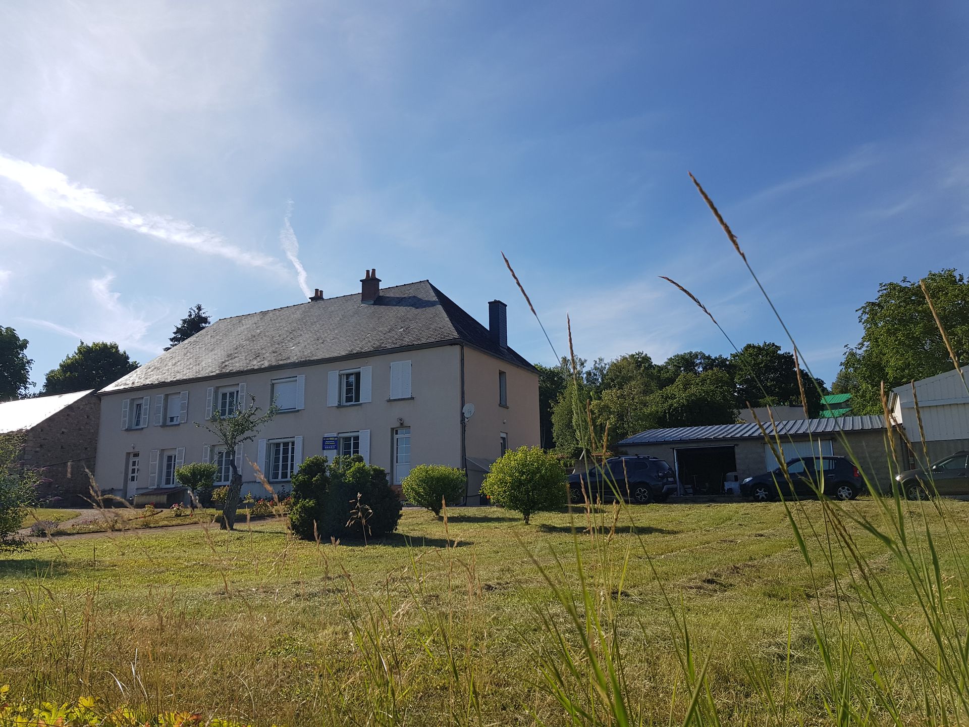 Une maison pâle au toit sombre se dresse derrière un champ herbeux sous un ciel d'un bleu éclatant, avec un garage sur la droite.