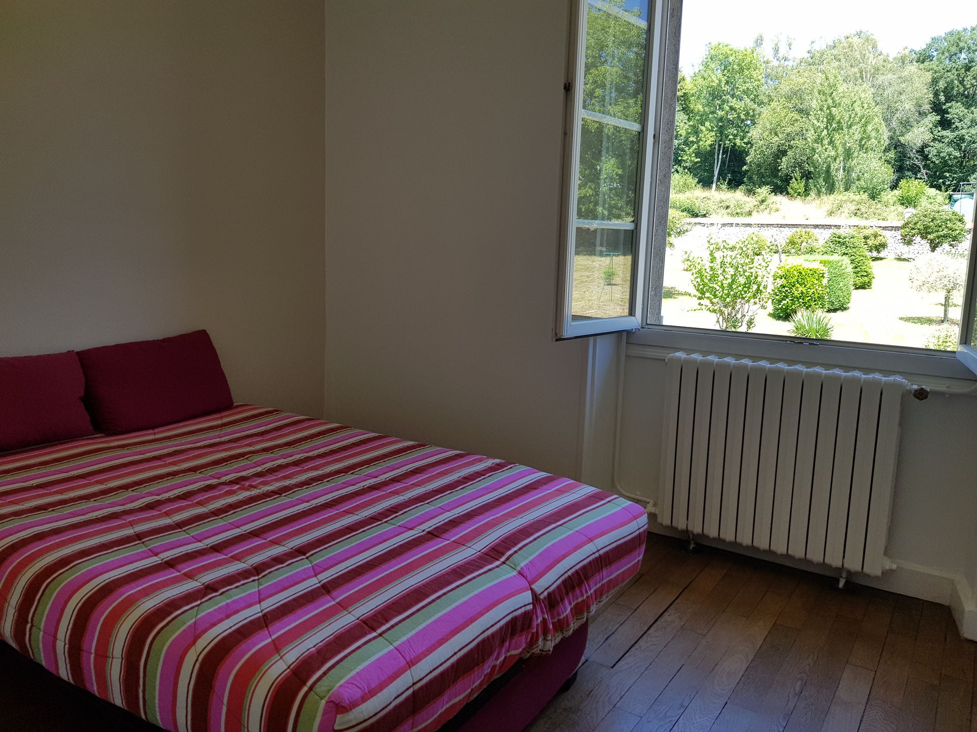 Une chambre avec un lit doté d'une couette à rayures roses et de deux oreillers foncés, près d'une fenêtre donnant sur un jardin.