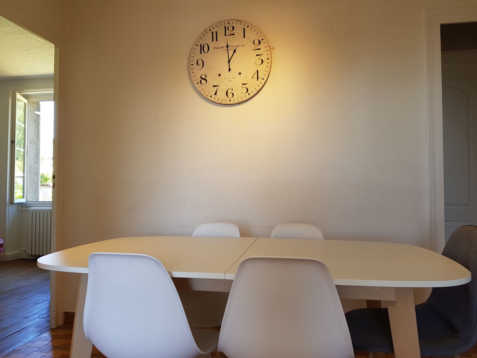 Dans une pièce où est fixée une grande horloge ronde sur un mur nu, se trouve une table à manger beige avec des chaises blanches.