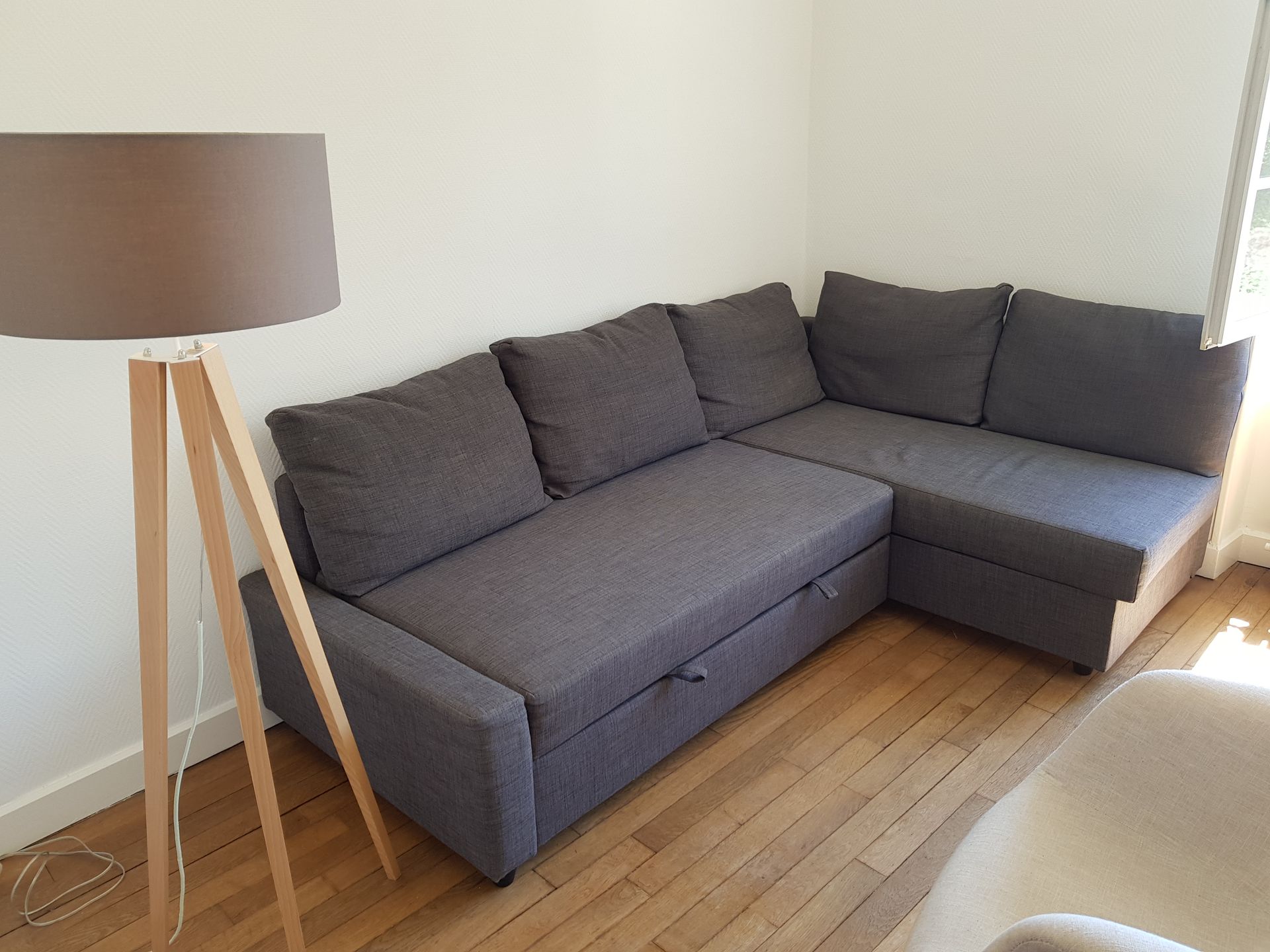 Un canapé d'angle gris est placé sur un parquet clair, à côté d'un lampadaire trépied à abat-jour marron.