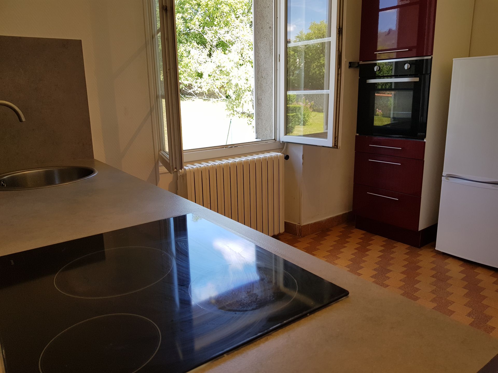 Une cuisine avec un plan de travail, une plaque de cuisson électrique, un évier, un radiateur près d'une fenêtre ouverte, un four encastré et un réfrigérateur.