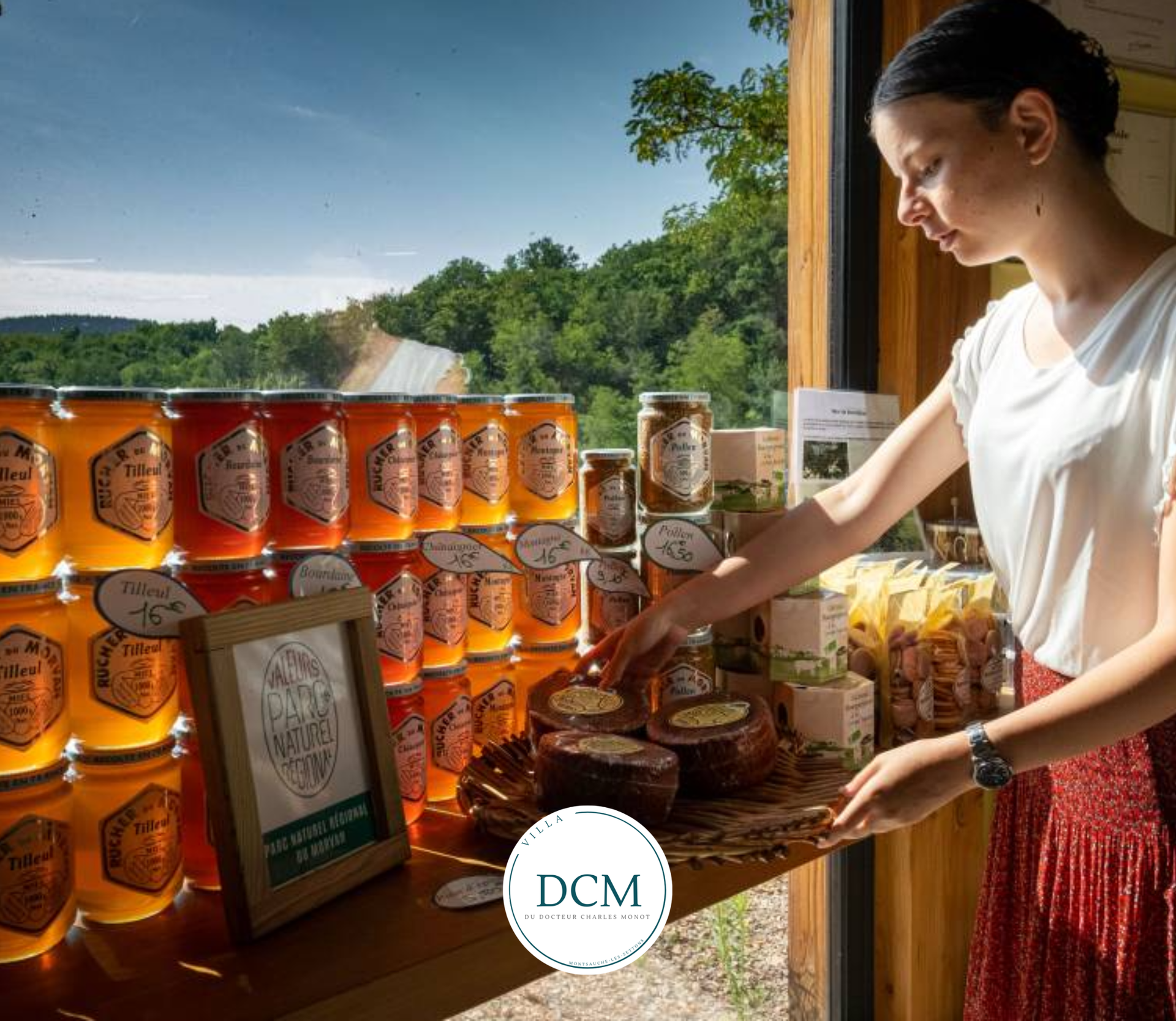 Une personne dispose des pots de miel et des produits emballés sur une table devant une fenêtre donnant sur un paysage boisé pittoresque.