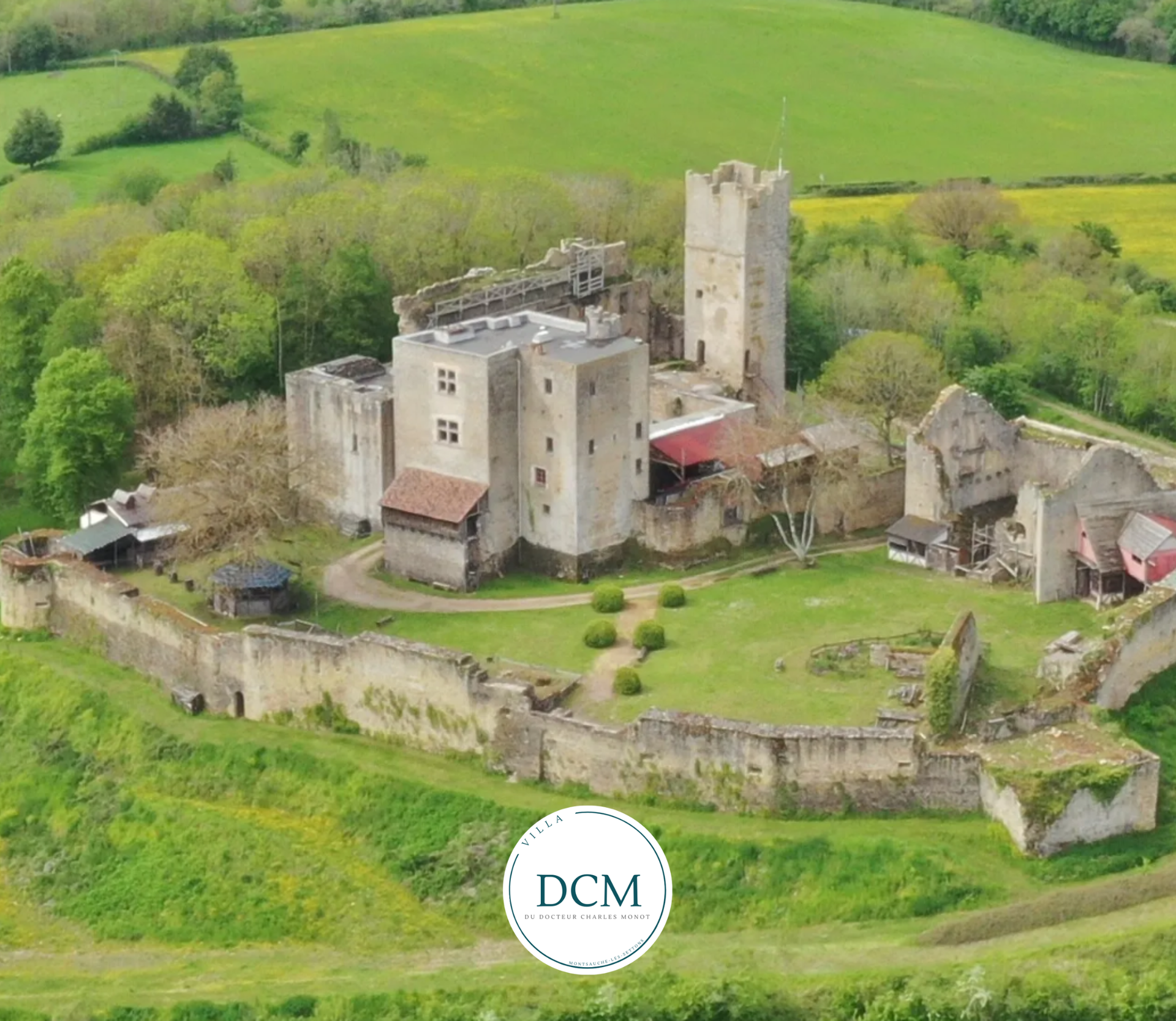 Vue aérienne des ruines d'un château en pierre entouré de champs verdoyants et d'arbres, avec une tour imposante et une cour intérieure.