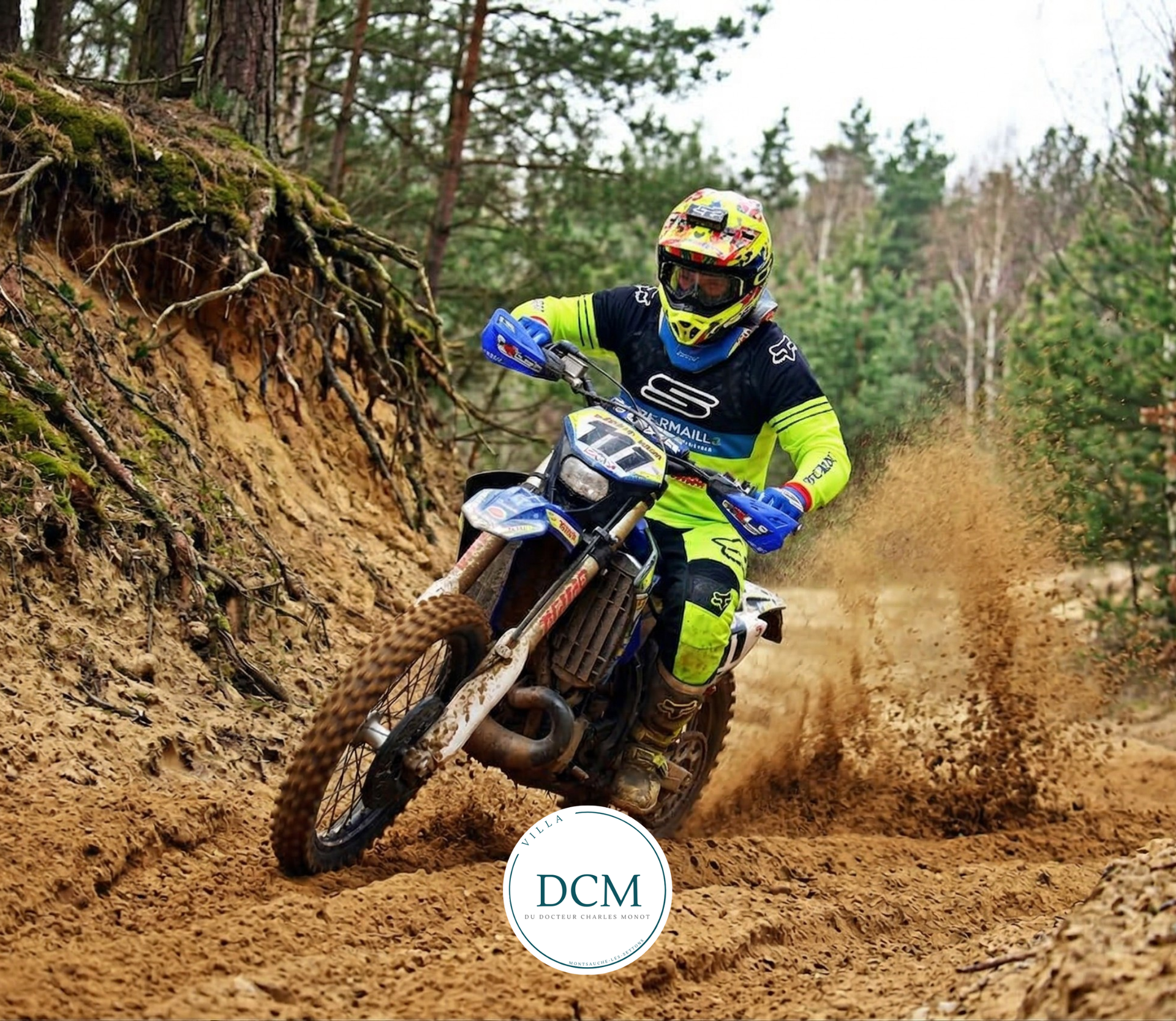 Un motard en maillot jaune et noir conduit une moto tout-terrain bleue sur un chemin forestier sablonneux, soulevant un nuage de poussière.