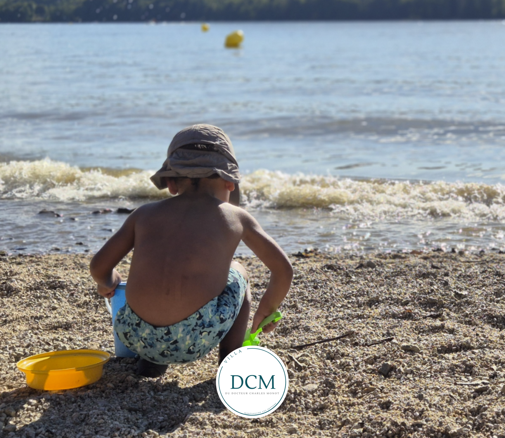 Une personne portant un chapeau de soleil et un maillot de bain à motifs joue avec un seau jaune sur une plage rocheuse au bord de l'eau.