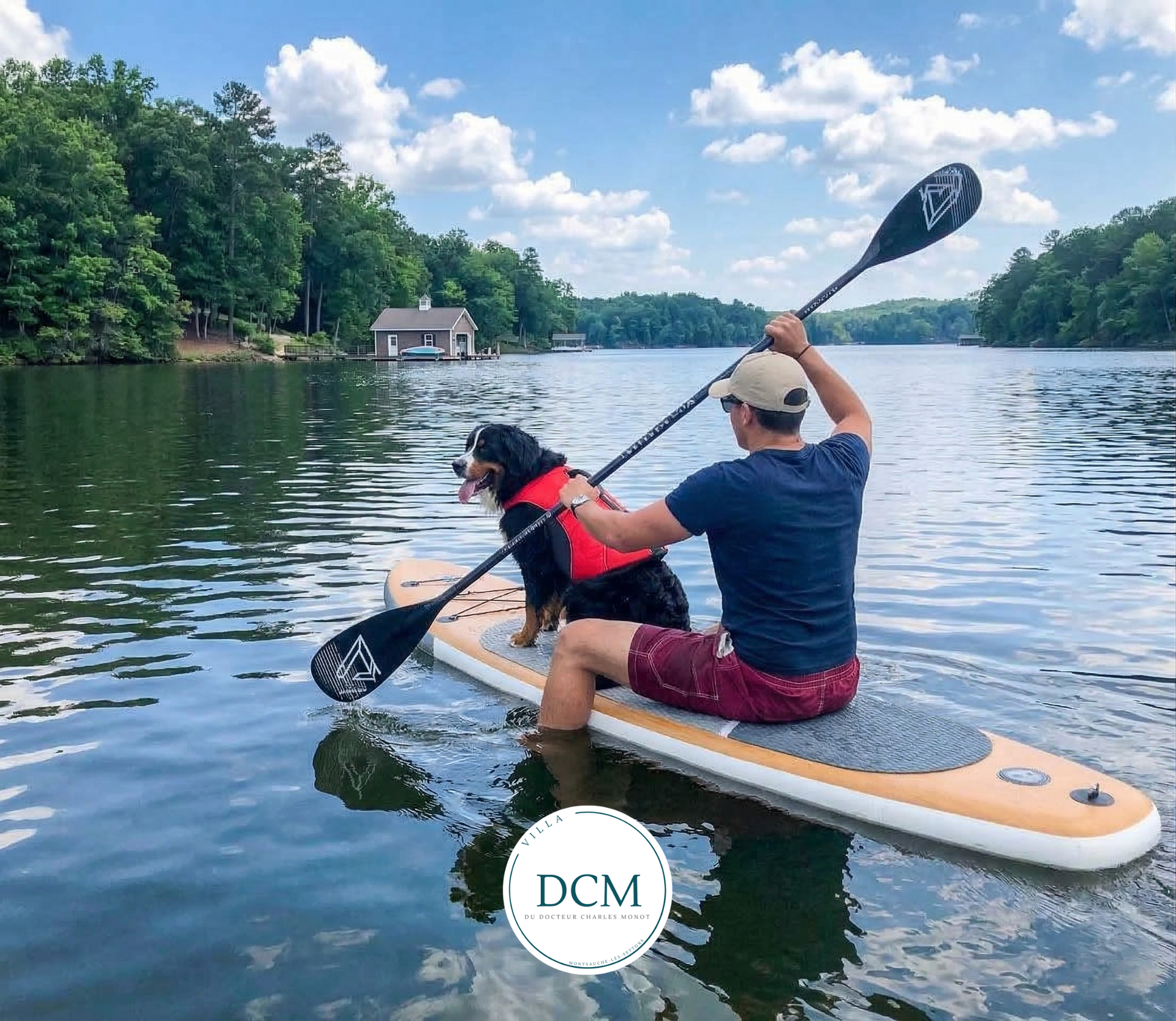 Une personne fait du paddle avec un chien portant un gilet de sauvetage rouge sur un lac calme et ensoleillé, entouré d'arbres.
