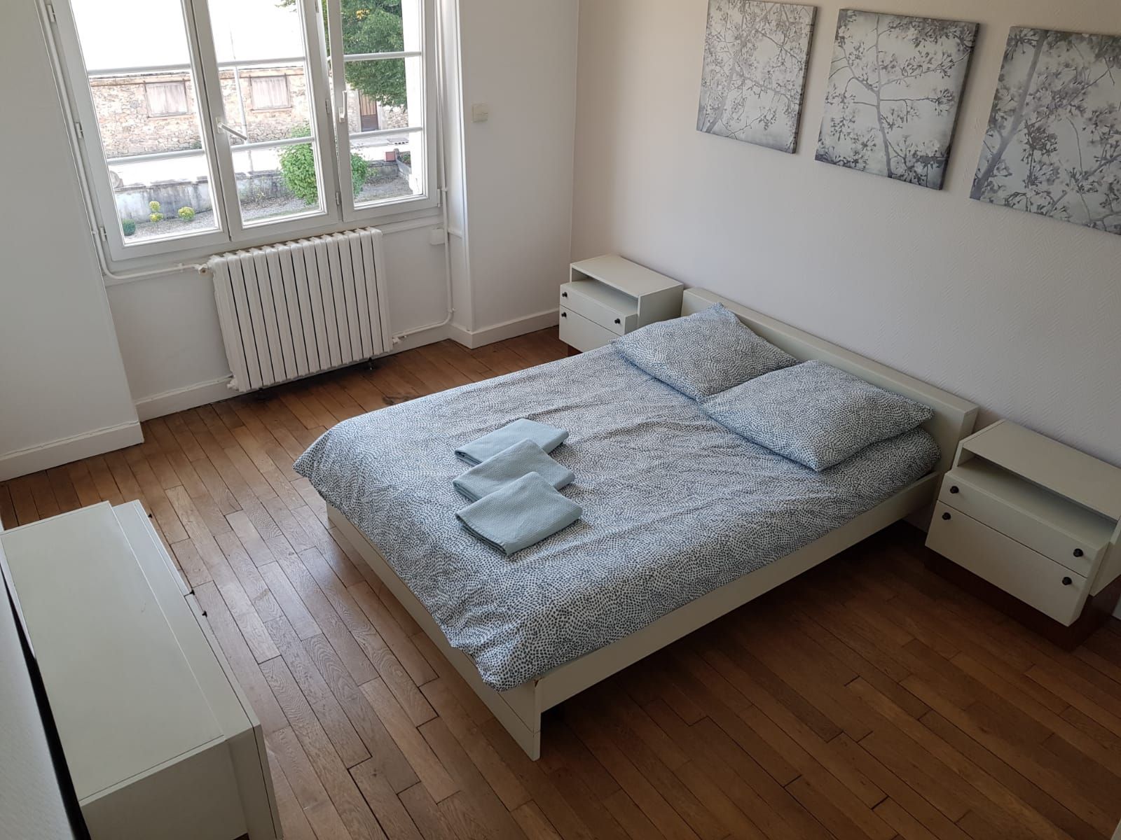 Une chambre lumineuse et bien rangée avec un lit double, du linge de lit à motifs clairs, du parquet, deux tables de chevet et trois reproductions murales.