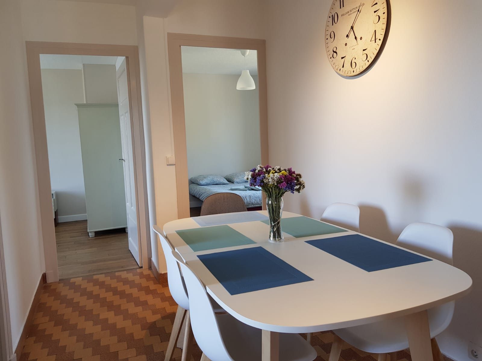 Une table à manger dressée pour quatre personnes avec des sets de table colorés et un vase de fleurs, dans une pièce avec une horloge murale et deux portes.