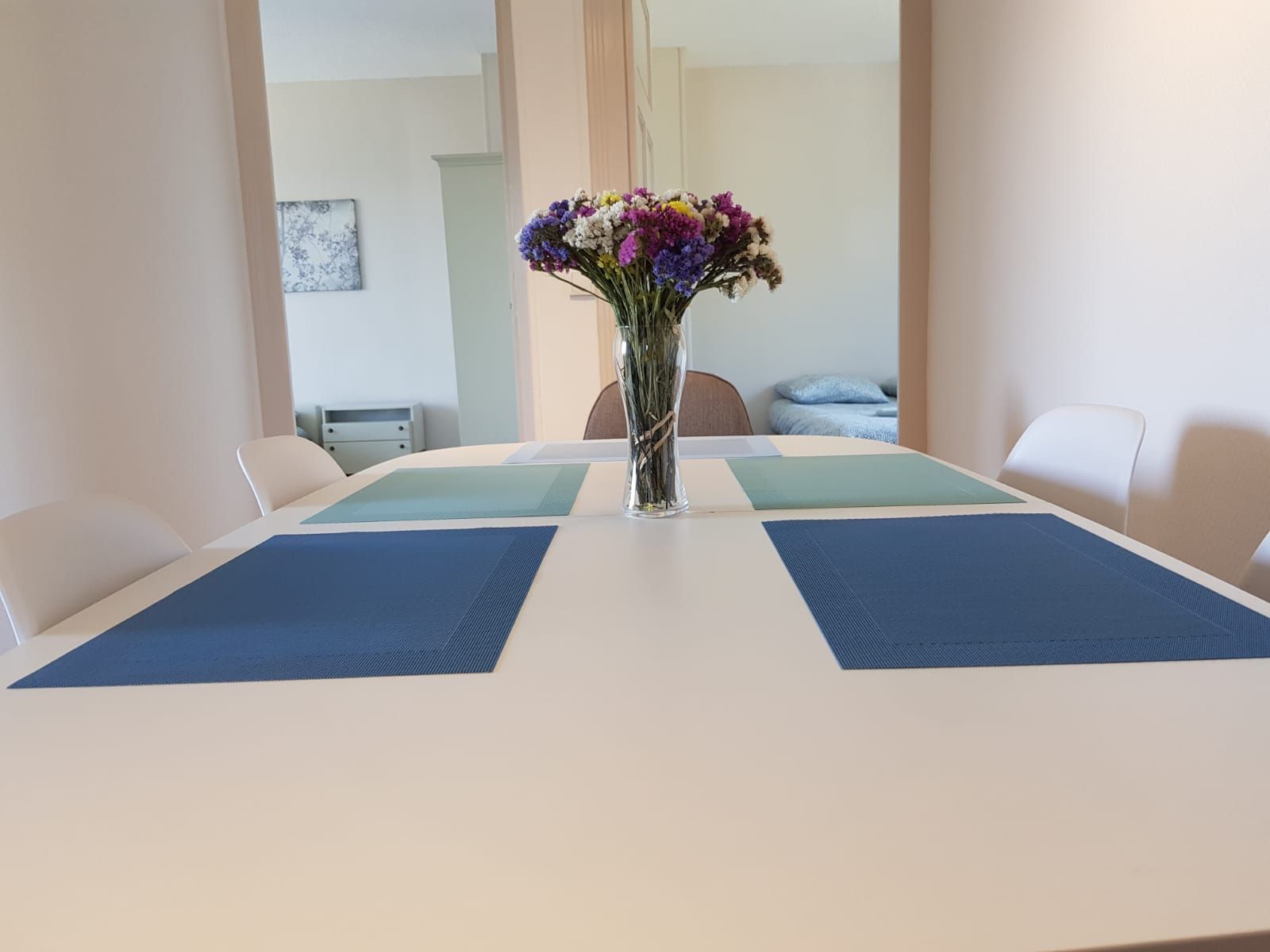Une table à manger avec des sets de table bleus et un vase de fleurs, face à une porte ouverte donnant sur une autre pièce.
