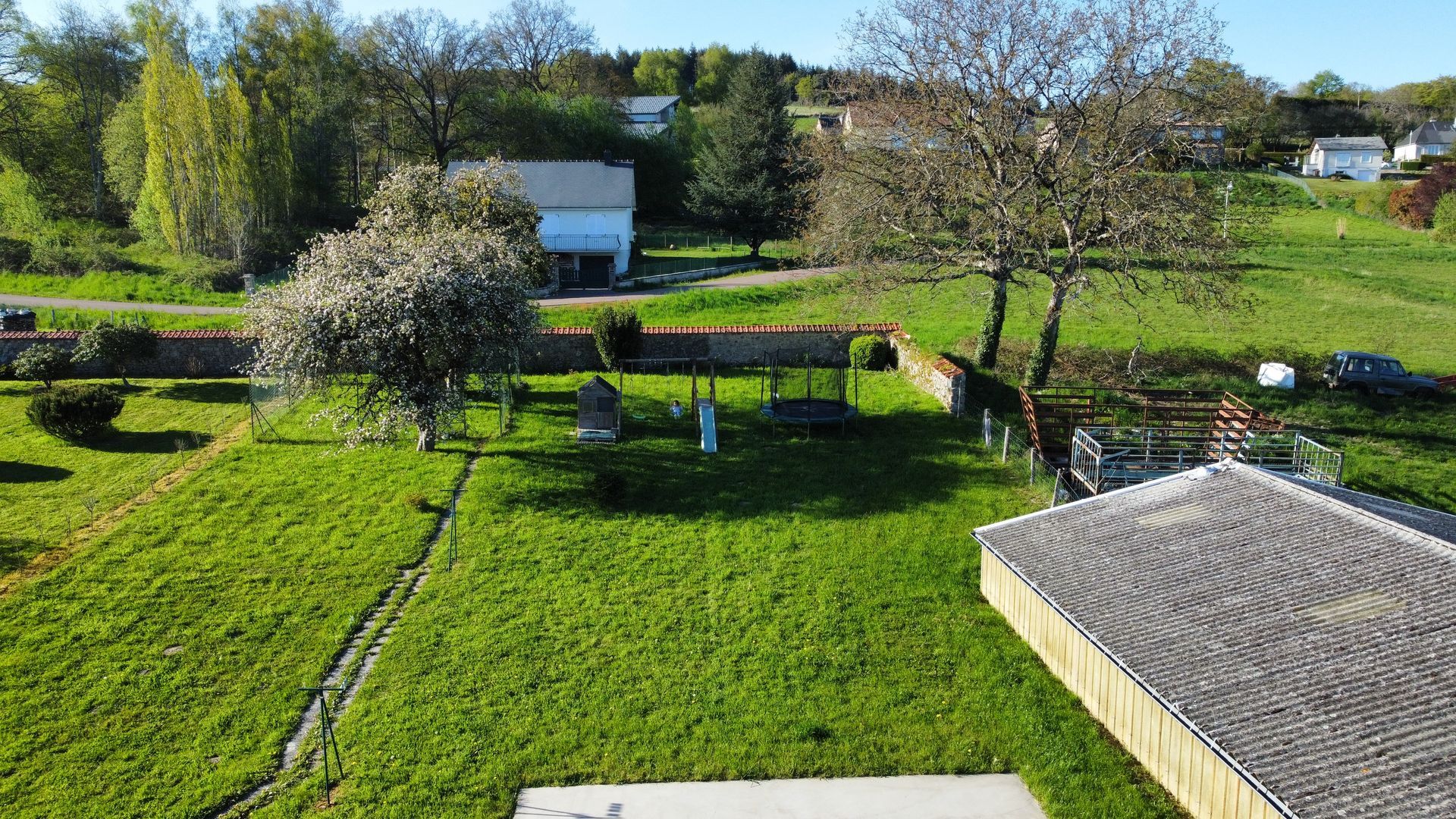 Un jardin de campagne avec une pelouse verte, un arbre en fleurs, un mur de pierre et un abri de jardin au toit en métal, sous un ciel bleu.