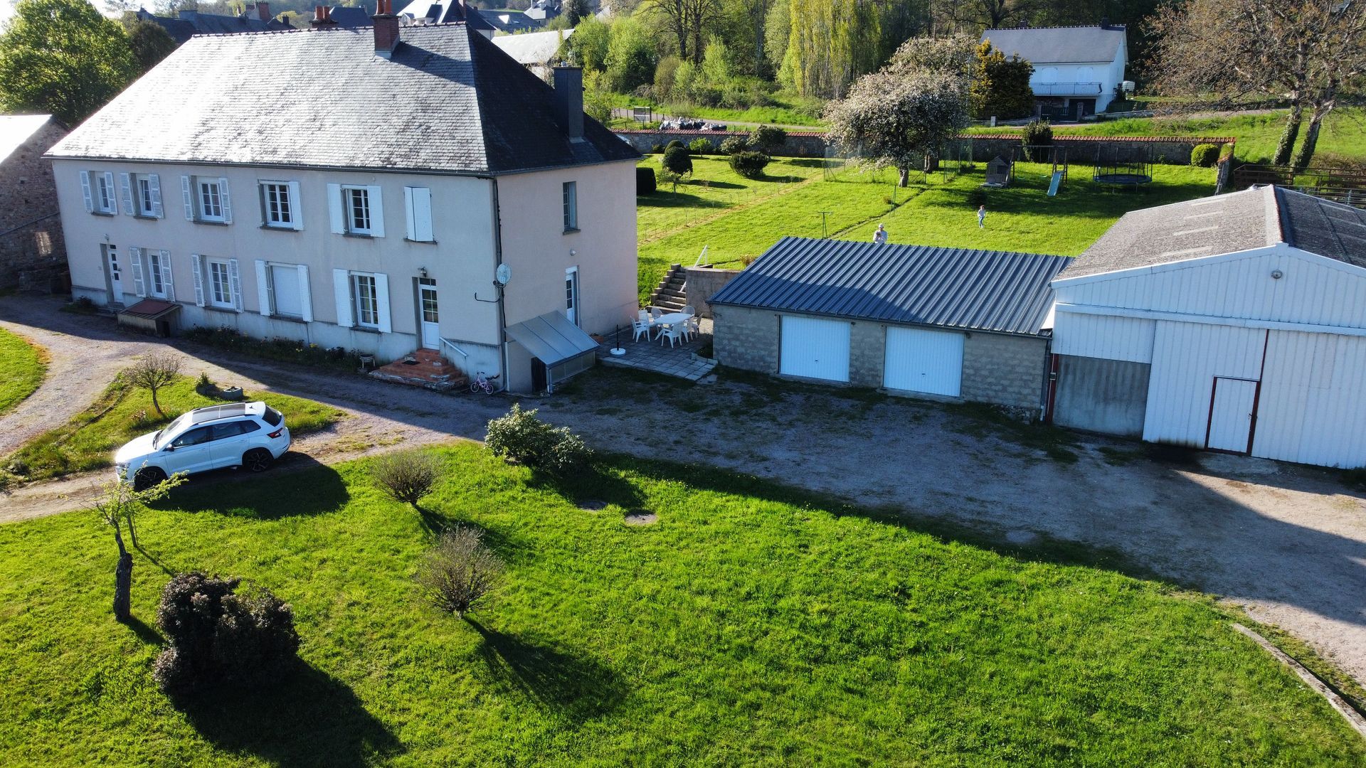 Vue aérienne d'une ferme beige à deux étages avec un toit gris, une voiture blanche et des garages adjacents sur un terrain verdoyant.