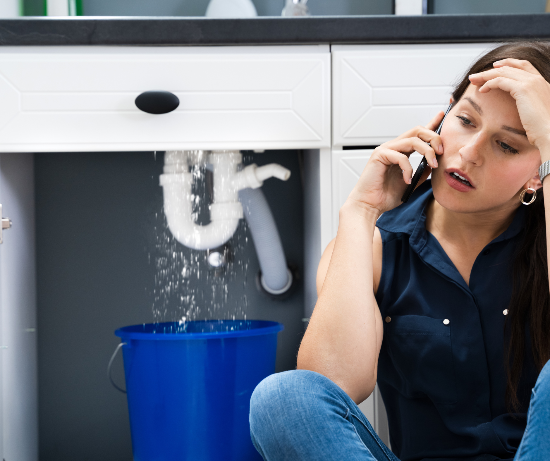 Woman on phone, looking stressed, next to a leaking sink, with a blue bucket collecting water.