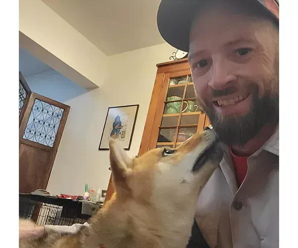 Man smiling, being nuzzled by a light brown dog indoors.