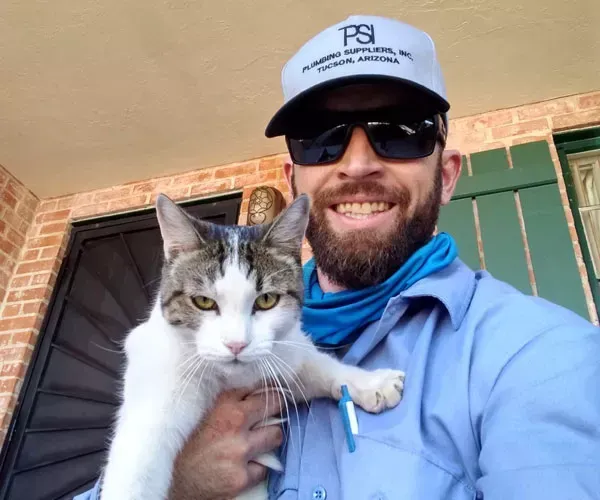 Man in cap and sunglasses holding a cat; they are smiling outdoors.