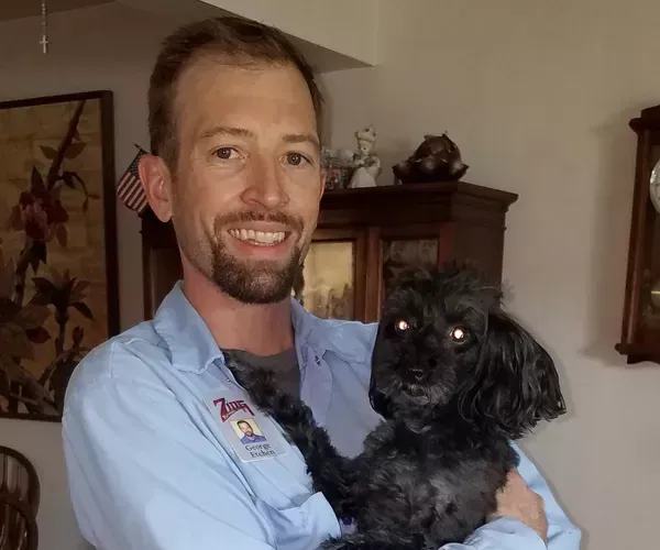 Man in light blue shirt holding a small, black dog, smiling. Indoors, with furniture in the background.