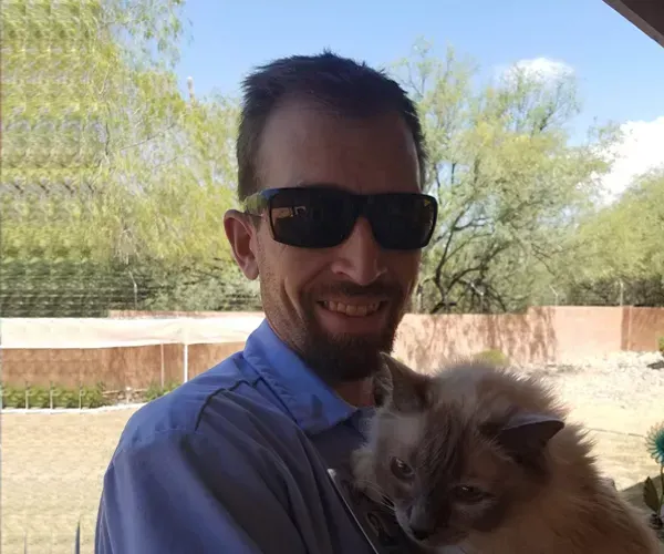 Man with sunglasses smiles, holding fluffy cat outdoors near trees and a fence.