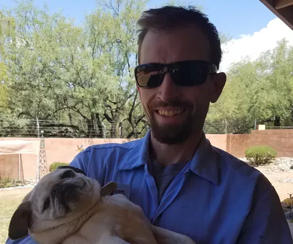 Man in sunglasses holds a small bulldog puppy, smiling outdoors in front of a tree.