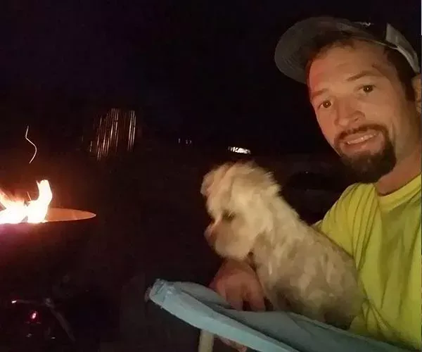 Man smiling, holding a small white dog by a campfire at night.