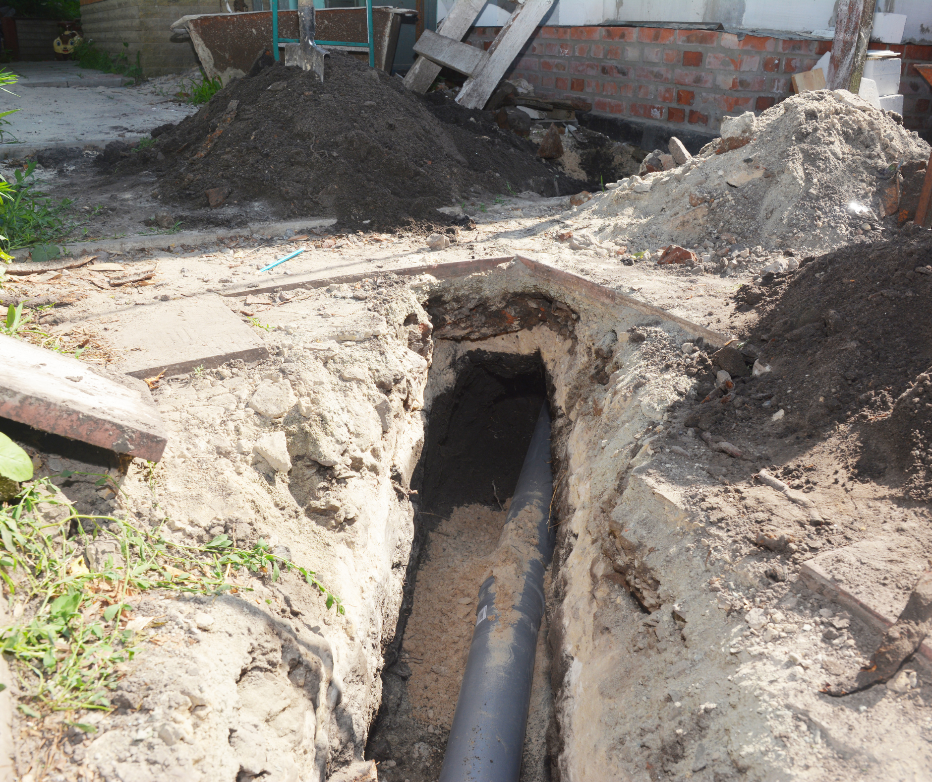 Trench dug with a pipe exposed; dirt and debris nearby; brick building in background.