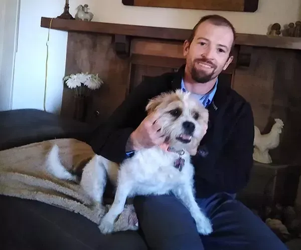 Man with a beard sits on a couch, petting a small, fluffy white and brown dog.