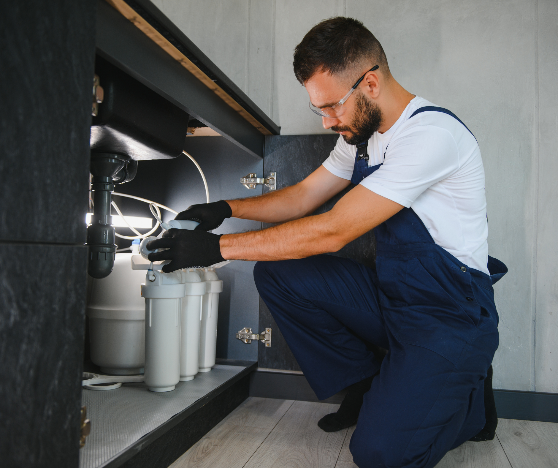 Plumber kneeling under a sink, working on a water filtration system; black gloves and navy jumpsuit.