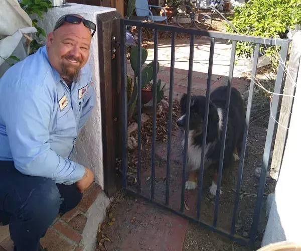 Man kneels next to a gate; a black and white dog is inside. The man is smiling.