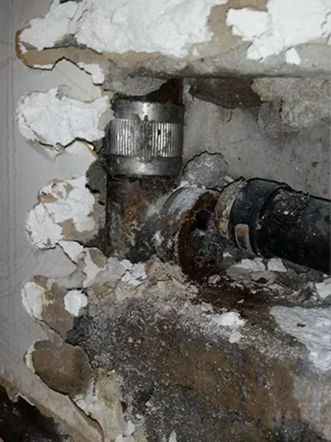 Pipes and fittings, possibly leaking, in a damaged wall, with white coating and dark stains.