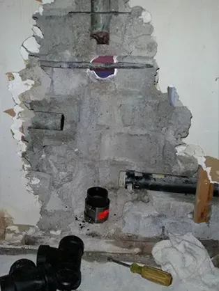 Exposed plumbing and pipes in a wall with damaged concrete and plaster. Black pipes, repair supplies visible.