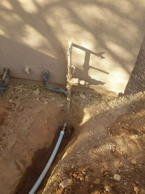 A buried water pipe in a trench leads to a metal valve, near a wall and a shadow of a wrench.