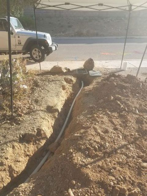 Trench with buried pipes, dirt mound beside a road. White Jeep parked behind a work tent.