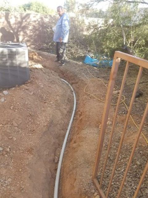Man standing near a trench with white pipe. Brown dirt surrounds. Rusty fence and a water tank visible.