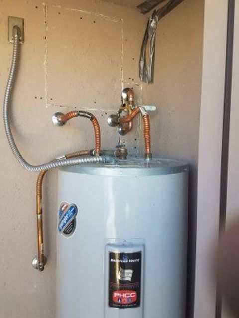 White water heater with copper pipes, electrical conduit, and a pressure relief valve against a beige wall.