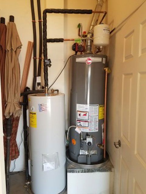 Two water heaters, one gray and one white, in a small utility room. Black pipes overhead.