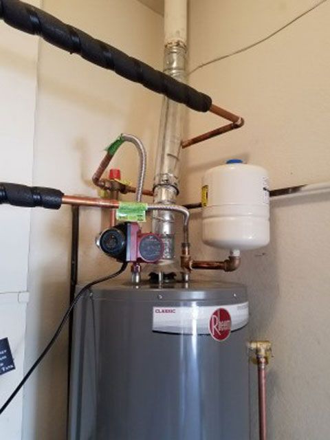 A water heater with copper pipes, a pump, and a white expansion tank in a room.