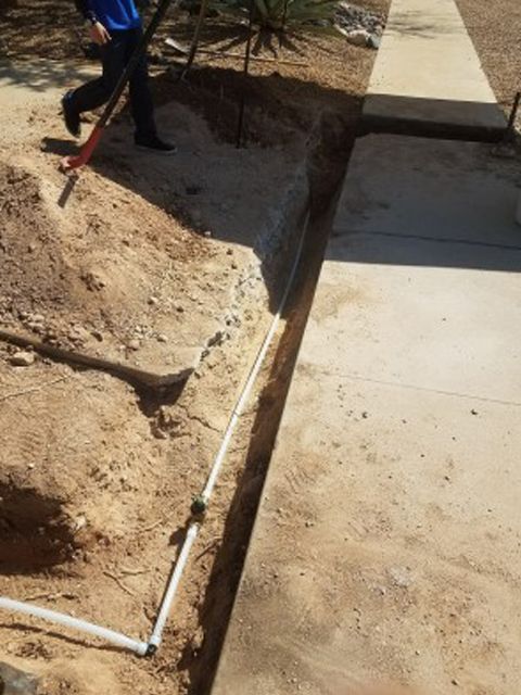 Person digging a trench next to a concrete walkway, white pipe visible in the trench.
