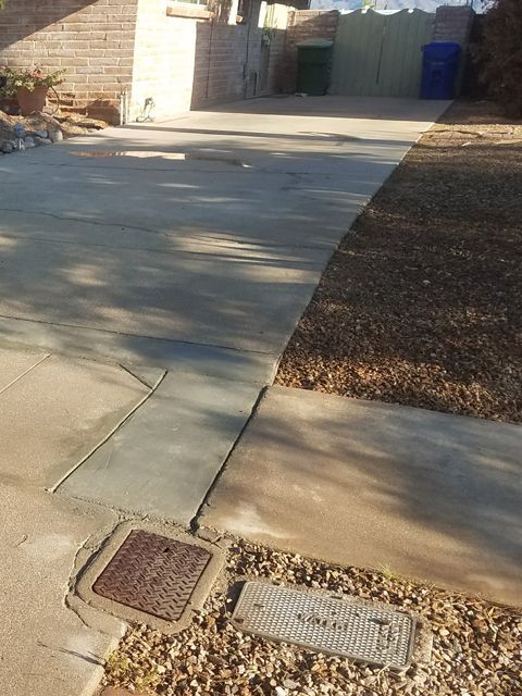 Concrete driveway with a utility access cover. Green and blue trash bins sit in the background.
