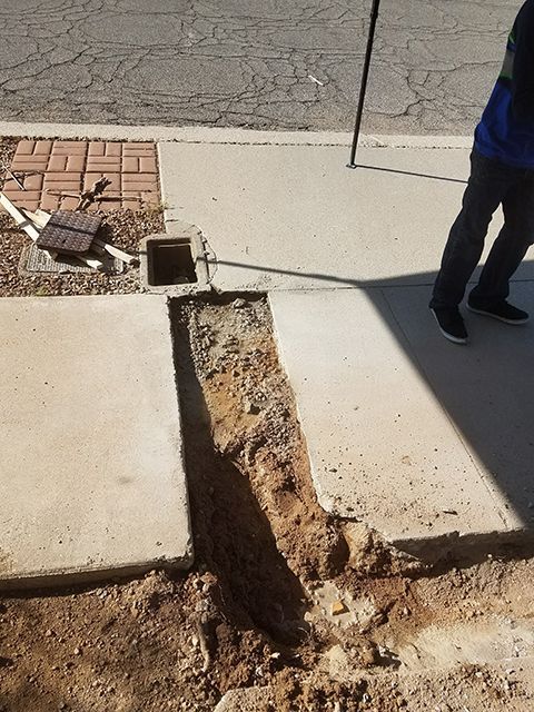 Trench cut into sidewalk, person standing nearby. Dirt exposed, brick and pipe visible. Sunlight casts shadows.