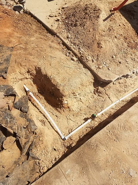 A trench in dirt with white PVC pipes laid inside.  Two mounds of dirt and a concrete surface surround.