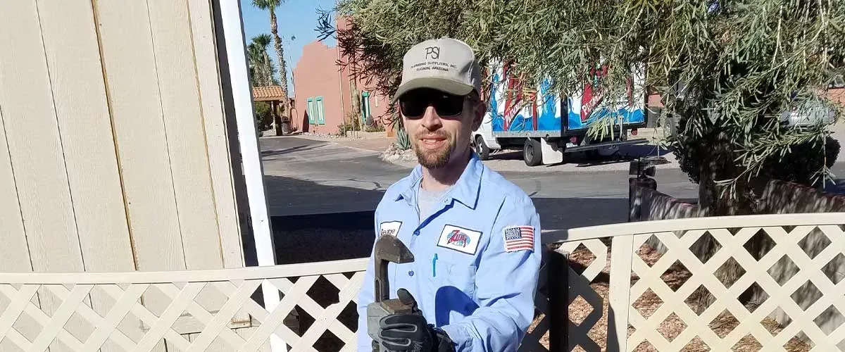 A man in a blue uniform and sunglasses holds a tool, standing in front of a white lattice fence.
