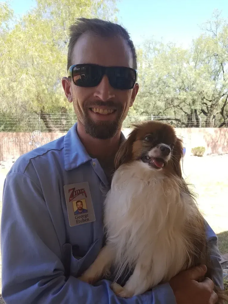 Man in sunglasses holding a small, fluffy dog outdoors, both smiling.