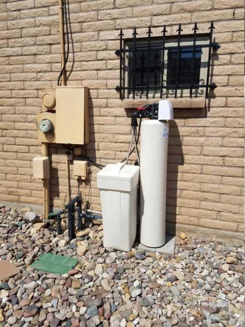 Exterior view of a beige brick building with utility boxes, a water softener, and a barred window.