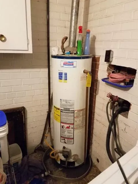 A water heater in a basement corner, next to white brick wall.  It has bottles on top and exposed wires.