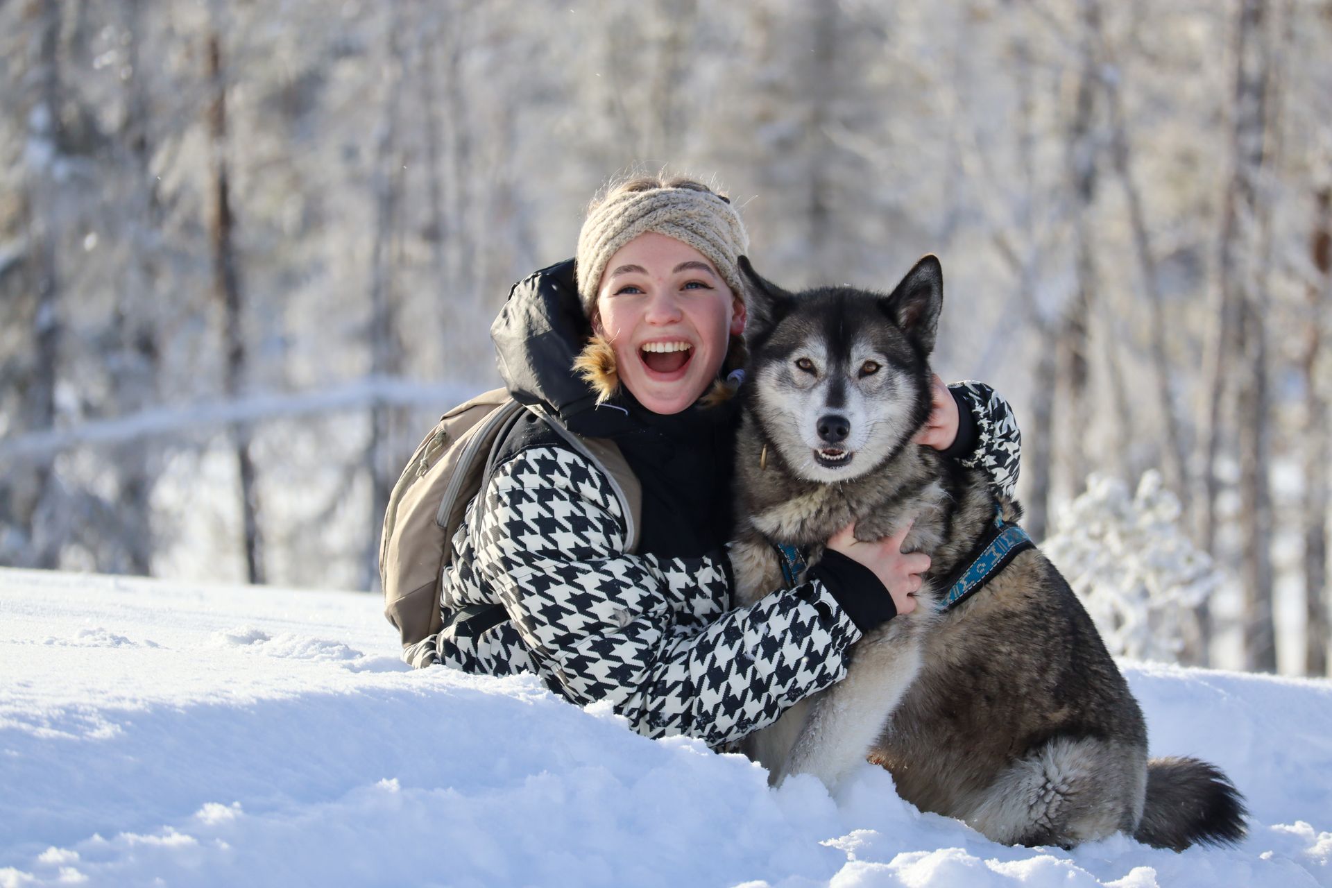 Een vrouw in een winterjas omhelst een husky in een besneeuwd bos. Ze lachen allebei vrolijk.