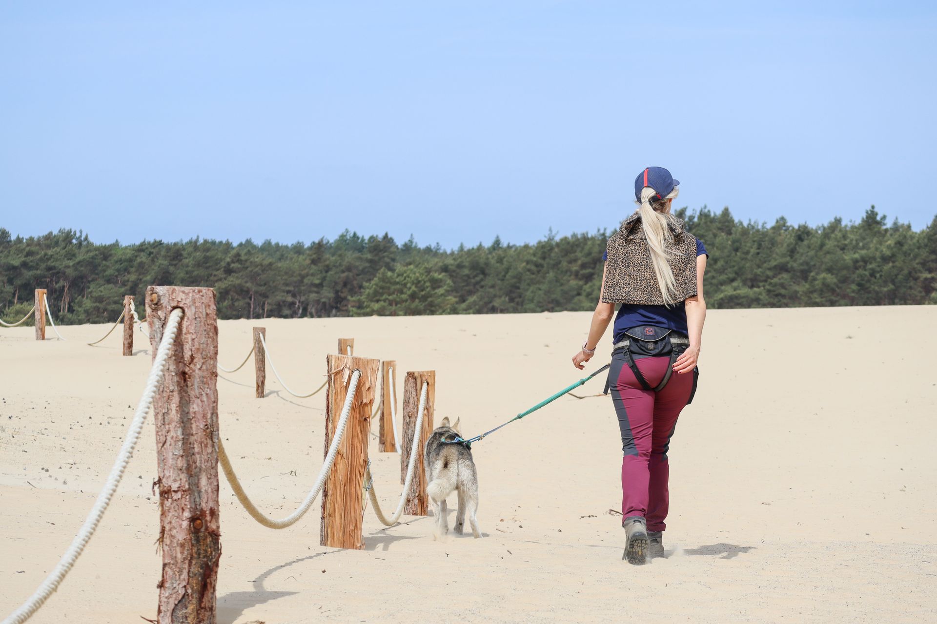 Een vrouw loopt met haar hond op een zandpad met een touwbarrière, met bomen op de achtergrond en een blauwe lucht.