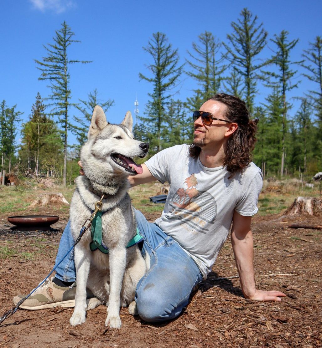 Een man met lang haar en een zonnebril knielt naast een husky in het bos. Hij lacht en ze kijken allebei omhoog.