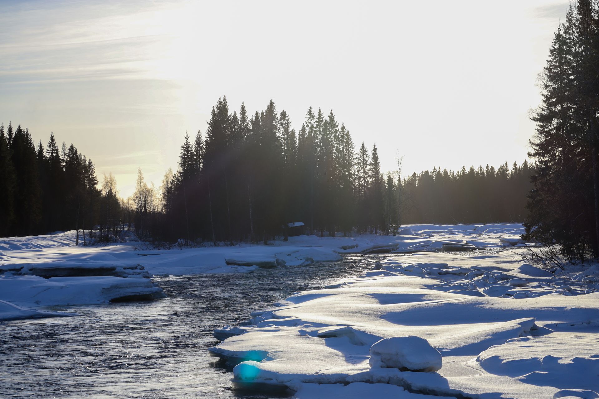 Besneeuwde rivier met bomen; zonlicht dat weerkaatst op het water en het ijs.