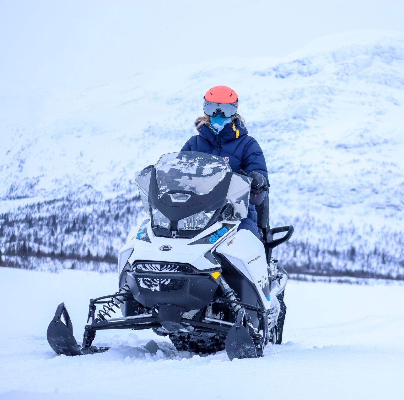 Persoon die op een witte sneeuwscooter rijdt in een besneeuwd landschap, met een blauwe jas, helm en gezichtsbedekking.