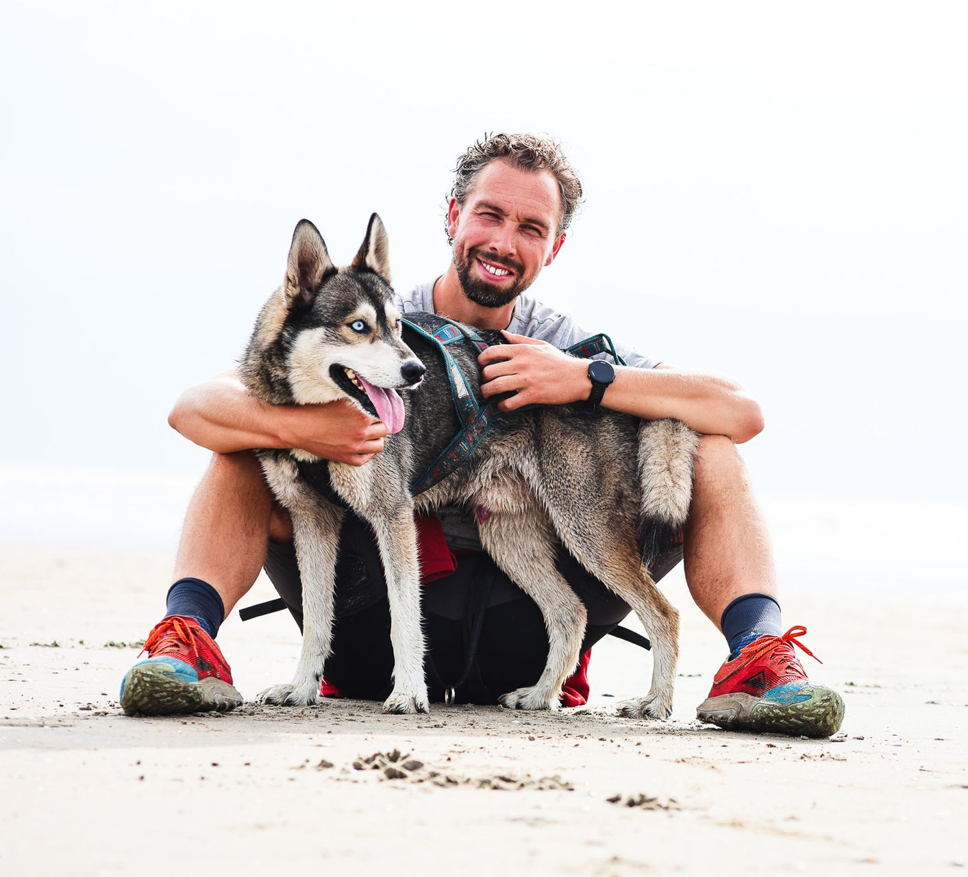Man zit op het strand en omhelst twee husky's.