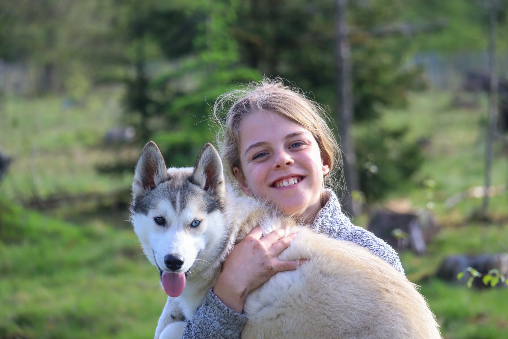 Een lachend meisje knuffelt buiten een husky. Groene bomen en gras op de achtergrond.