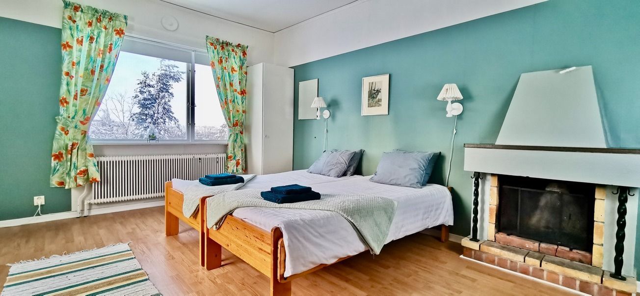 Slaapkamer met twee eenpersoonsbedden, een open haard en een raam met uitzicht op een besneeuwd tafereel. Turquoise muren, gordijnen met bloemenmotief en een houten vloer.